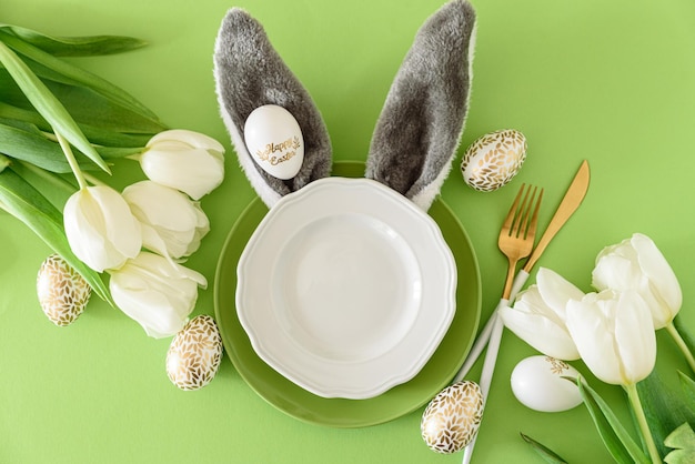 Empty plate decorated bunny ears white tulips and Easter eggs on green background