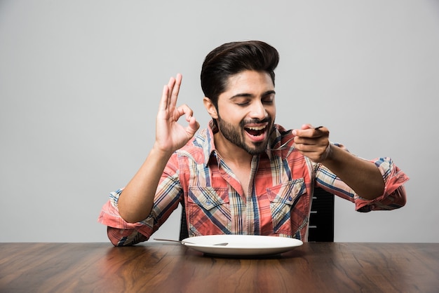 Empty plate and Indian man with beard holding spoon and fork, wearing checkered shirt and sitting at table