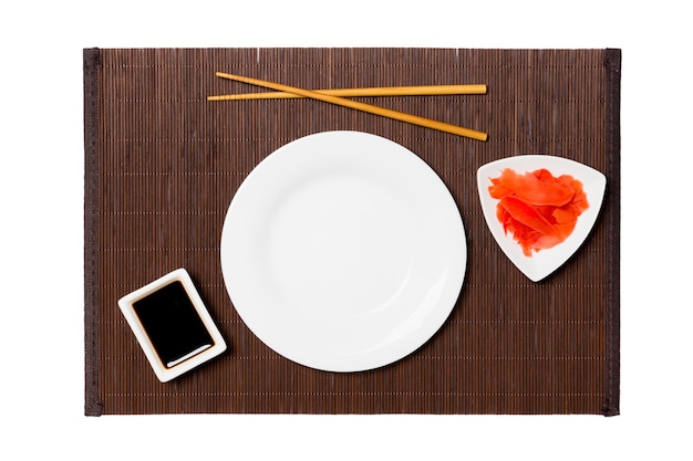 Empty round white plate with chopsticks for sushi, ginger and soy sauce on dark bamboo mat background. Top view with copy space for you design.