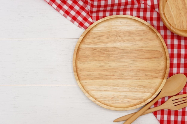 Empty round wooden plate with spoon fork and red gingham tablecloth on white wooden table