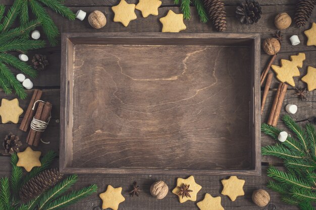 Empty rustic tray surrounded by ginger biscuits, fir branches and cones, cinnamon and walnuts. Christmas food background.