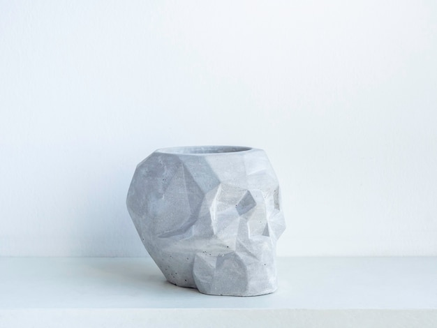 Empty skull shape concrete plant pot on white wooden shelf isolated on white wall. Small modern DIY cement planter trendy decoration.