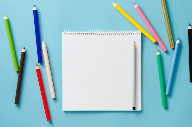 Empty square notebook and set of colorful pencils on blue background. Blank paper.