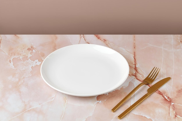 Empty white blank plate with gold fork and knife on beige pink marble background Minimalist mock up