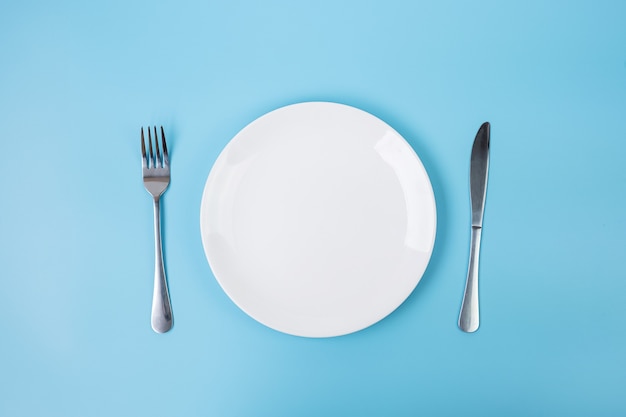 Empty white ceramics plate with knife and fork on blue background