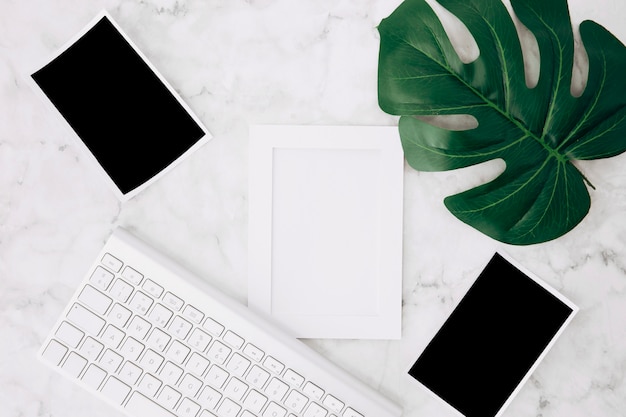 An empty white frame and instant photos with green monstera leaf and keyboard on desk