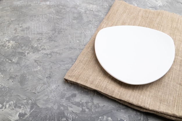 Empty white plate on gray concrete background and linen textile. Side view,