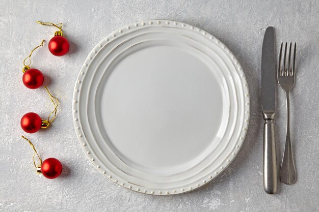 Empty white plate red Christmas balls and cutlery on a gray stone table New Year's menuu