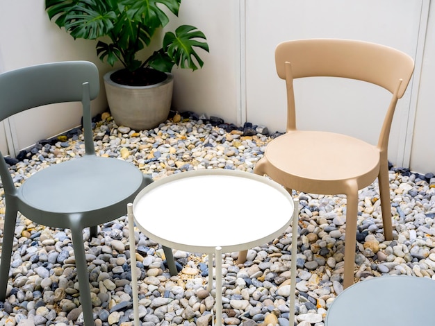 Empty white round shape side table near the empty green and yellow pastel colors chairs seats on the gravel ground floor near the green leaves in concrete pot in small room minimal style interior