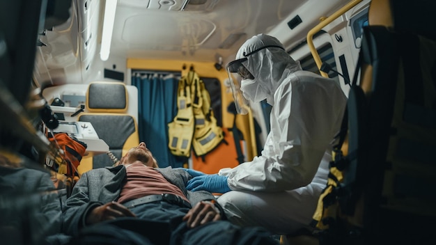 EMS Paramedic in Disposable Coverall Suit Comforting Injured Patient on the Way to Hospital Emergency Medical Care Assistant Puts His Hand on Vinctims Shoulder in a Friendly Way in an Ambulance