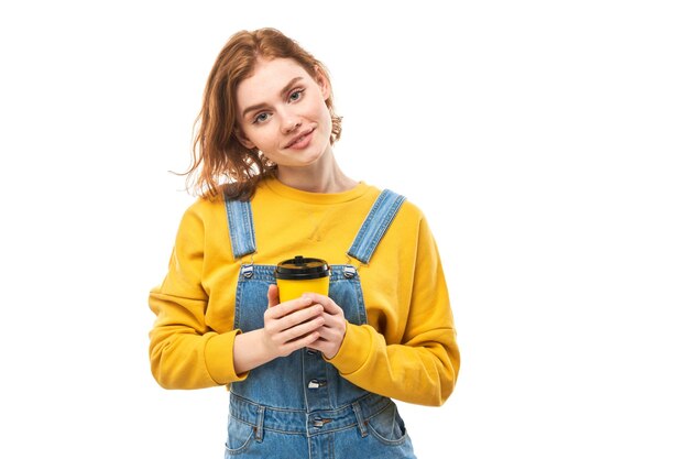 Energetic redhead girl holding and drinking yellow cup of takeaway coffee smiling and enjoying drink isolated on white background