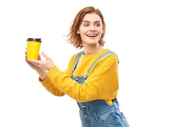 Energetic redhead girl holding and drinking yellow cup of takeaway coffee smiling and enjoying drink isolated on white background