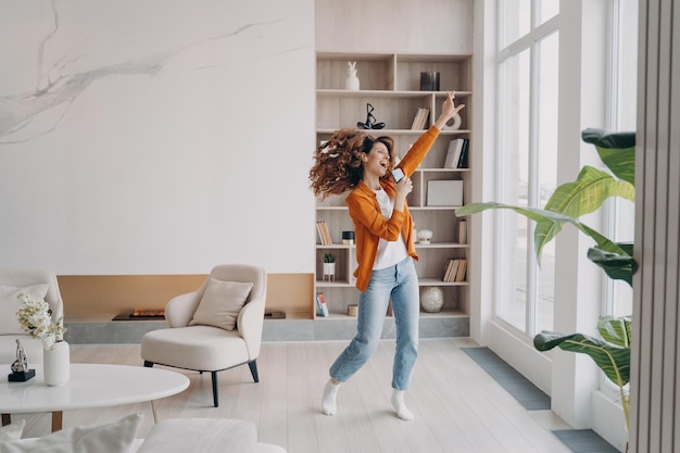 Energetic woman holding smartphone having fun dances to music rejoicing with good news at home