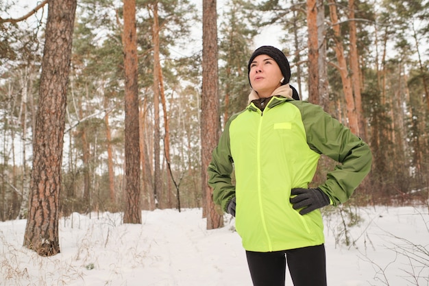 Energetic young woman in green jacket holding hands on hips and inhaling frosty air in forest while hiking alone