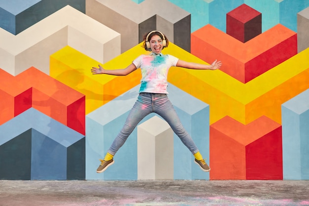 Energetic young woman with headphones jumping near colorful wall