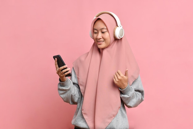 Energized happy muslim woman dances joyfully enjoys favourite music wears stereo headphones isolated on pink background expresses positive mood