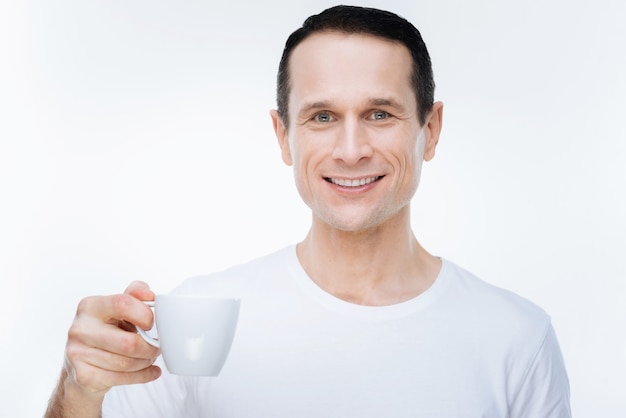 Energizing drink. Cheerful positive delighted man smiling and holding a cup of coffee while looking at you