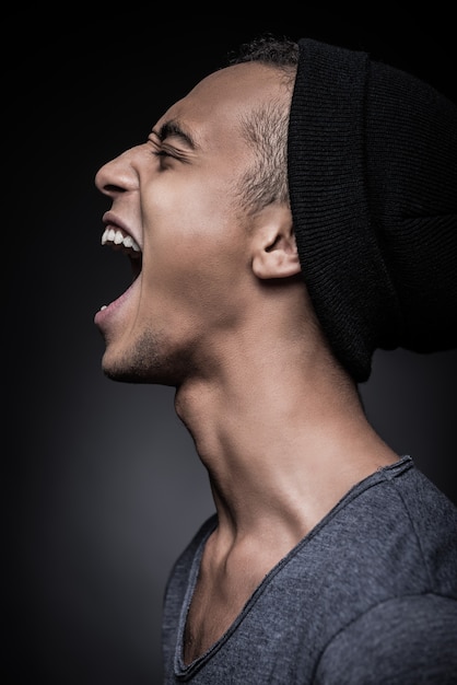 Energy inside him. Side view of furious young African man in beanie keeping eyes closed and mouth open while standing against black background