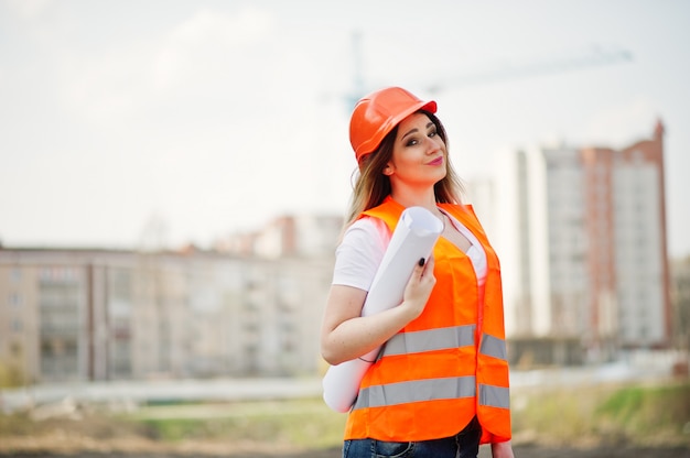 Engineer builder woman in uniform waistcoat and orange protective helmet hold business paper against new buildings with crane. 