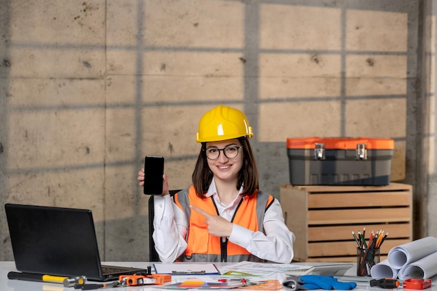 Engineer cute young smart brunette girl civil worker in helmet and vest with phone