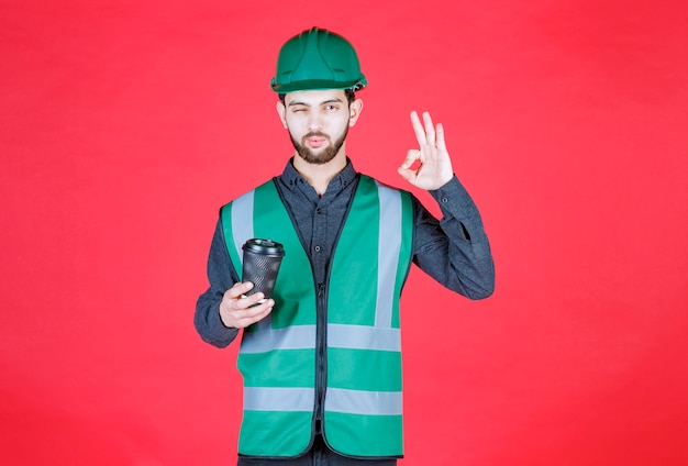 Engineer in green uniform and helmet holding a black disposable cup of coffee and enjoying the taste. 