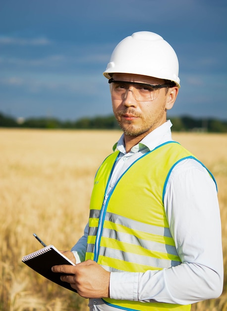 Engineer in a helmet, goggles and a yellow vest, a hand on the helmet holding a notepad, a folder