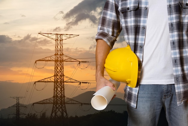 Engineer man construction hand holding yellow helmet hardhat on electric pole at power plant sunset background Worker helmet Civil Construction Engineering Close up engineer hand hold safety helmet