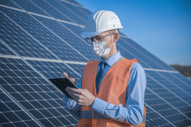 Engineer, man in uniform and mask, helmet glasses and work jacket on a of solar panels at solar station