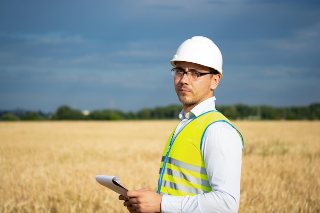 An engineer in a white helmet and a yellow vest stands with a folder in the field a farmer in the f