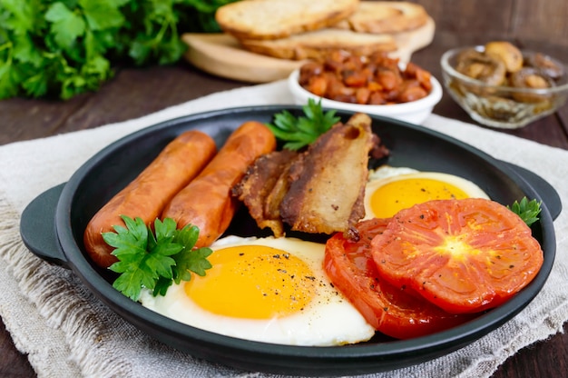 English breakfast: sausages, bacon, tomatoes, egg, beans in sauce, fried mushrooms, toast on a dark wooden table. Top view. Traditional classic food of England.