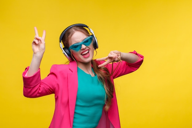 Enjoy listening to music. Young redhead woman in headphones listening music. Funny smiling girl in earphones and pink jacket dancing and singing on yellow background. Relaxation and stress management.