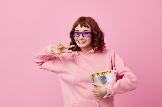 Enjoyed happy smiling cute redhead lady in pink hoodie sunglasses with popcorn posing isolated on over pink studio background Copy space Banner Fashion Cinema Movie concept Entertainment offer