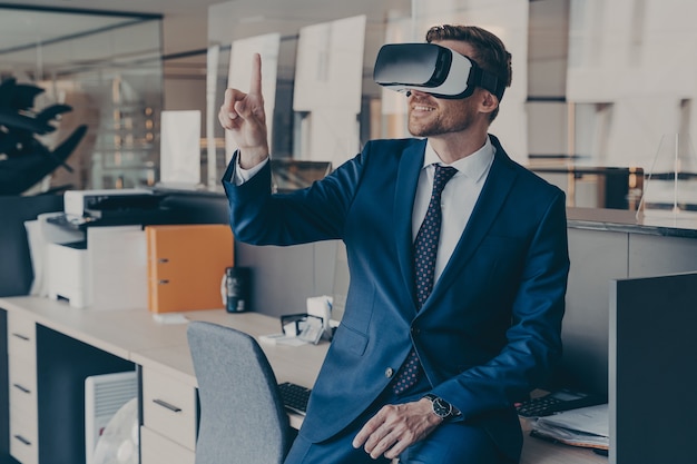 Enjoyed young businessman pointing with forefinger up in air with virtual reality glasses on head, while sitting on table in office with VR headset. Innovative gadget for business 3d visualizations