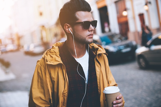 Enjoying city life. Handsome young man in headphones carrying coffee cup while walking along the city street