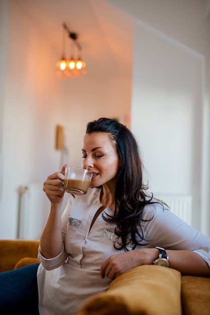 Enjoying coffee at home. Beautiful brunette drinking coffee, sitting on the sofa.