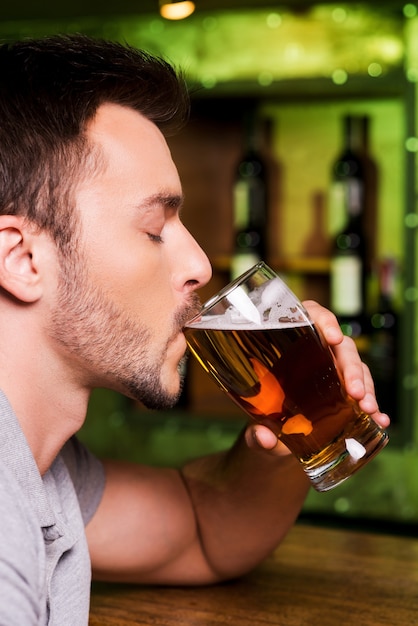 Enjoying cold and fresh beer. Side view of young man drinking beer while sitting at the bar counter