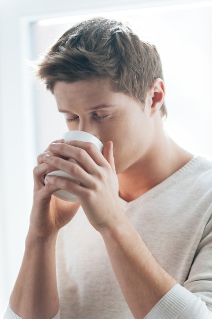 Enjoying fresh coffee. Handsome young man drinking coffee while standing against window