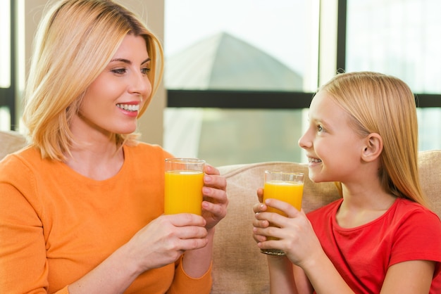 Enjoying fresh juice. Happy mother and daughter holding glasses with orange juice and looking at each other with smile while sitting on the couch together