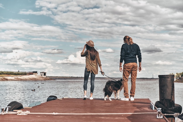 Enjoying great view. Full length rear view of beautiful young couple with dog standing on the wooden platform while spending time near the river