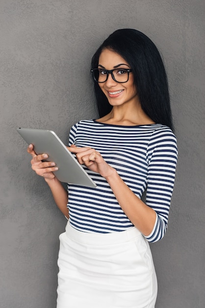 Enjoying her new tablet. Beautiful young African woman working on digital tablet and looking at camera with smile while standing against grey background