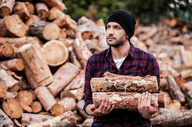 Enjoying his work with wood. Confident young forester holding logs and looking away while standing outdoors