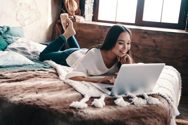 Enjoying lazy time at home. Beautiful young woman using laptop and smiling while lying on the bed at home