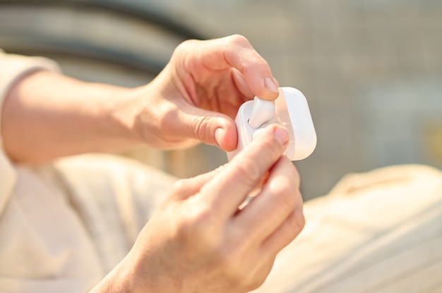 Enjoying moment. White beautiful case with modern wireless headphones in neat hands of woman illuminated by sunlight, without face