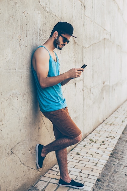 Enjoying music outdoors. Full length of handsome young man in headphones holding smart phone and looking at it while standing outdoors and leaning at the concrete wall