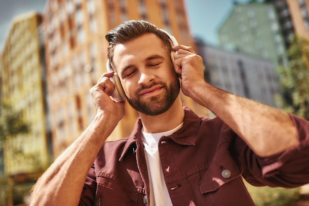 Enjoying my favorite song. Handsome young bearded man in headphones listening to the music and keeping eyes closed while standing on the street. Inspiration. Music concept. Sound