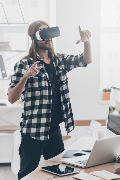 Enjoying new reality. Handsome young man with long hair in virtual reality headset gesturing while standing in creative office