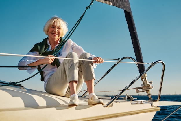 Enjoying sea trip happy senior woman sitting on the side of a sail boat or yacht deck floating in