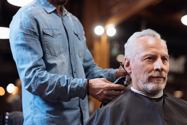 Enjoying service. Low angle of nice senior bearded man sitting while barber trimming his hair at barbershop.