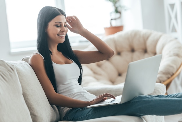 Enjoying small chat with friend. Attractive young woman typing something on laptop and smiling while relaxing on the couch at home