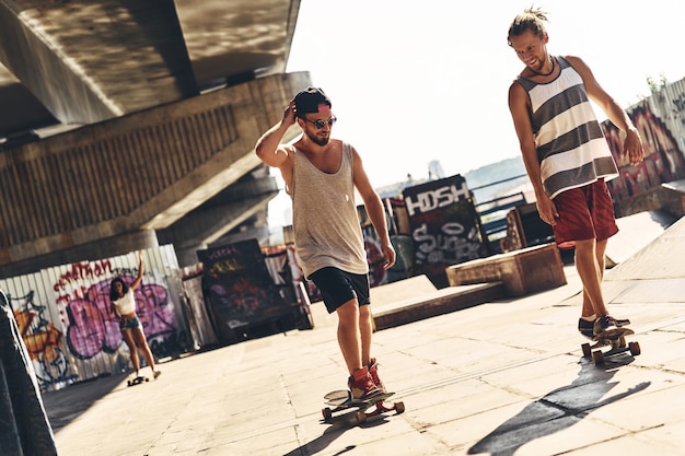 Enjoying sunny day. Full length of two handsome young men skateboarding while hanging out with their friends outdoors